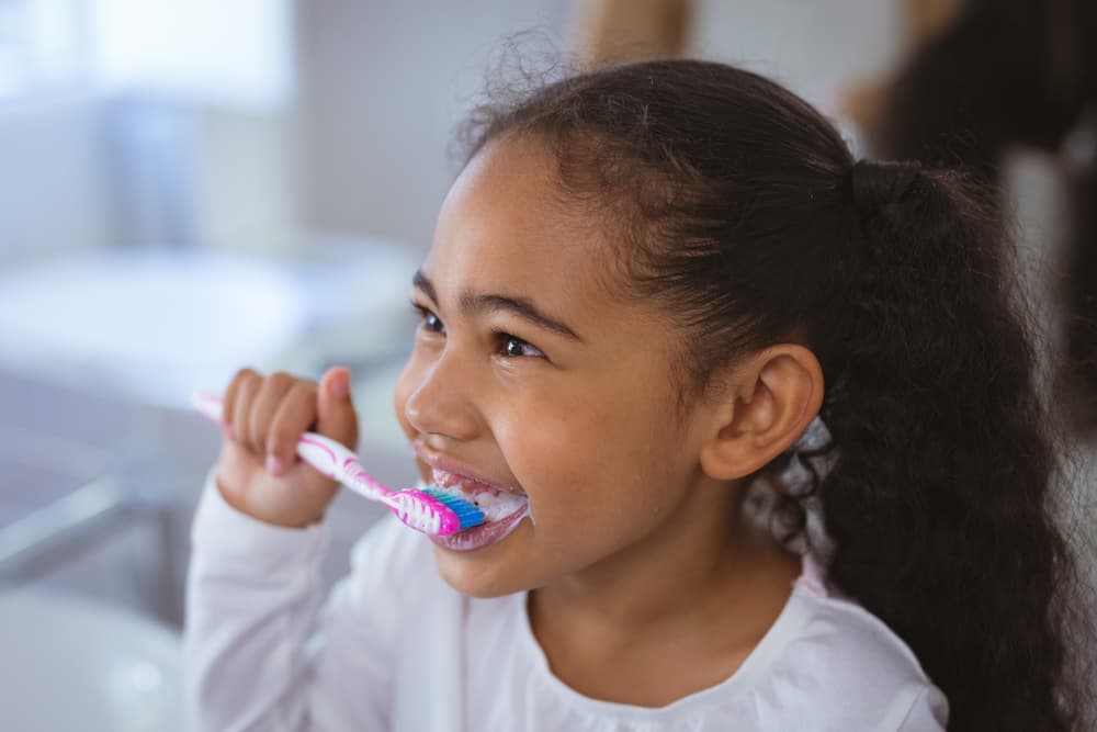 Smiling young girl brushing her teeth with a pink and blue toothbrush, emphasizing the importance of teaching proper oral hygiene techniques for daily maintenance.